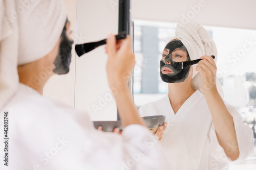 woman in a clay rejuvenating moisturizing face mask is smiling. Good disorder is positive self-love. Facial spa treatment at home in front of the mirror. Weekend morning routine. Girl young lifestyle