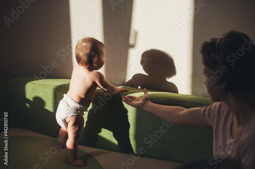 Adorable mother is helping her baby making her first steps holding her hand on a couch photo