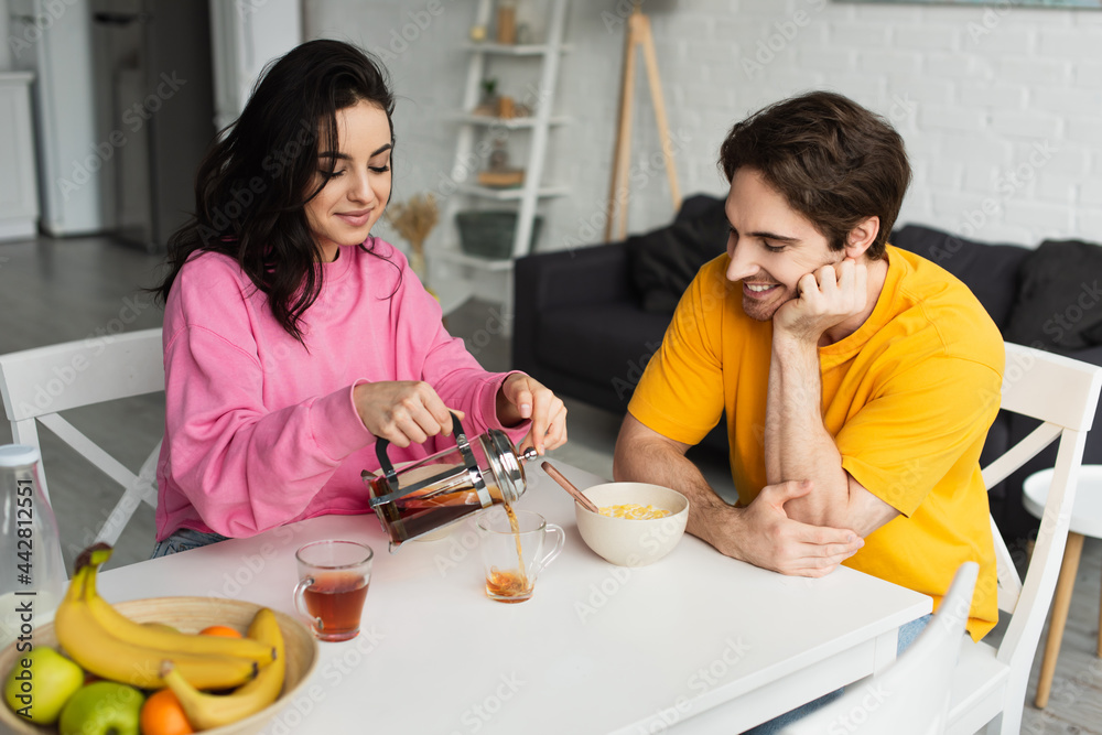 smiling young woman sitting at table near boyfriend with breakfast and pouring tea from french press into cup in living room