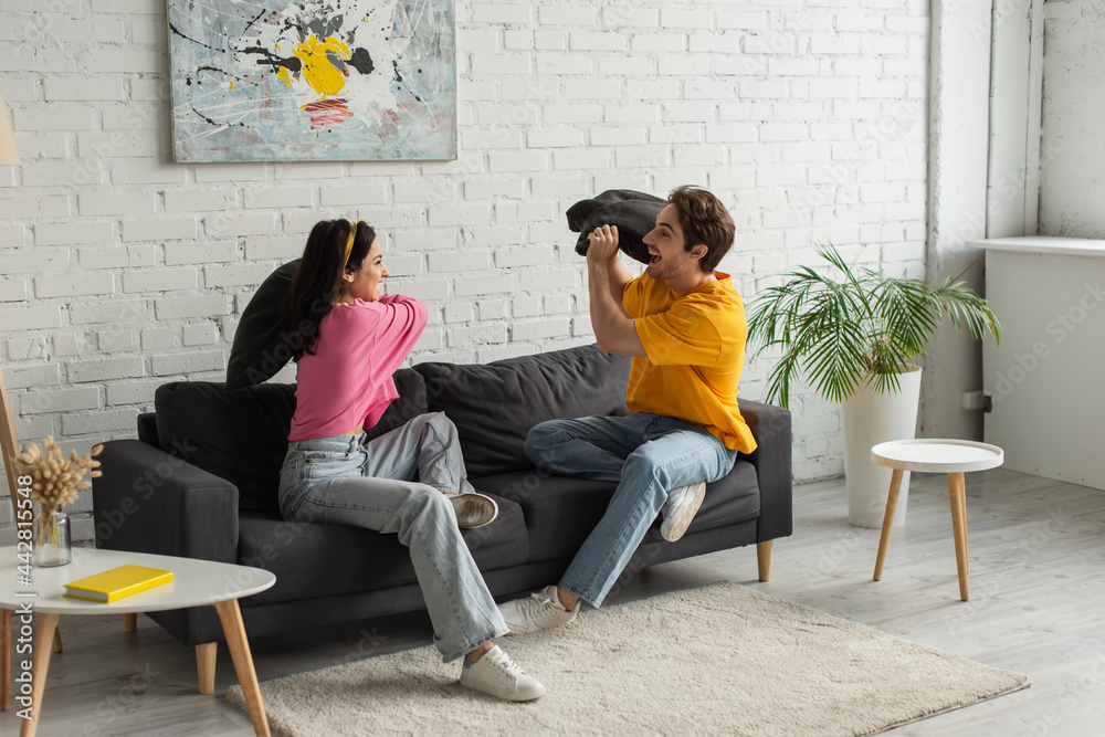 Fototapeta premium smiling young couple fighting with pillows on couch in living room