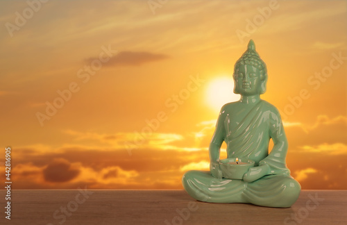 Beautiful ceramic Buddha sculpture with burning candle on wooden surface at sunset. Space for text