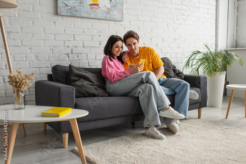 positive young couple sitting on couch and holding cellphone in living room