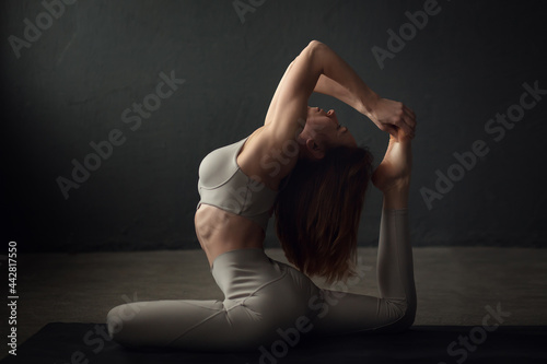 A slim beautifully lighted young  woman is doing One-footed King Pigeon yoga pose ose (Eka Pada Rajakapotasana) in a dark room. Image with selective focus and toning