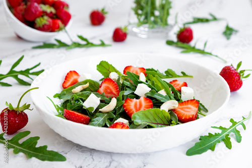 Summer salad of strawberry with arugula, spinach leaves, feta cheese and cashew nuts in white plate on marble table with salad leaves and fresh strawberry around. Strawberry salad recipe DIY. Step 4.