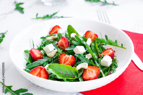 Salad of strawberry with arugula, spinach leaves, feta cheese, balsamic vinegar and cashew nuts with red napkin, salad leaves and fresh strawberry fruits around. Healthy food and raw food diet..