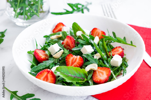 Vitamin salad of strawberry with arugula, young beet tops, feta cheese and cashew nuts in white plate with red napkin, salad leaves and fresh strawberry fruits around. Fruit detox and antioxidants.