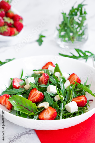 Vitamin salad of strawberry with arugula, young beet tops, feta cheese and cashew nuts in white plate with red napkin, salad leaves and fresh strawberry fruits around. Keto diet and paleo diet.