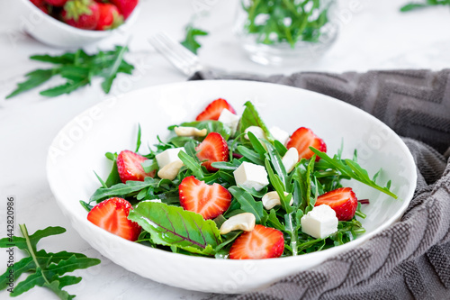 Vegan salad of strawberry with arugula, spinach, feta cheese and cashew nuts in white plate with gray towel, salad leaves and fresh strawberry fruits around. Healthy nutrition and vitamins concept.
