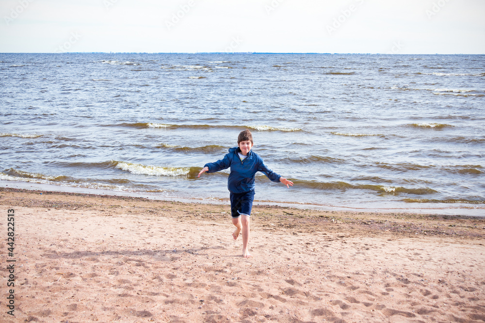 Happy boy in blue jacket and shorts barefooted running on sand on the beach, sunny day.