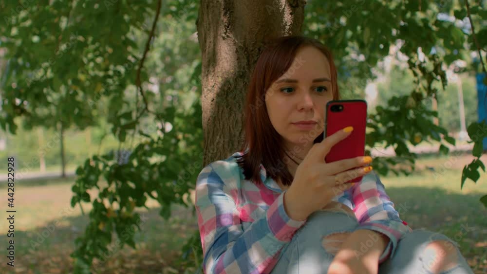 Close up of young woman rests with mobile phone, sitting near tree in park. Brunette browsing smartphone, sitting in shade near tree trunk in summertime Close up of young woman rests with mobile phone, sitting near tree in park. Brunette browsing smartphone, sitting in shade near tree trunk in summertime