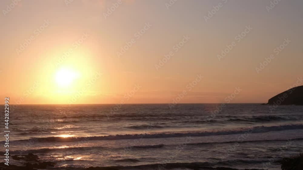 Colourful sunset over the ocean with low smooth cloud in Croyde, Devon, UK. A calm sea is revealed on a hazy evening as the time lapse zooms out. Cliffs and rocks look out to sea as the burning sun lo Colourful sunset over the ocean with low smooth cloud in Croyde, Devon, UK. A calm sea is revealed on a hazy evening as the time lapse zooms out. Cliffs and rocks look out to sea as the burning sun lo