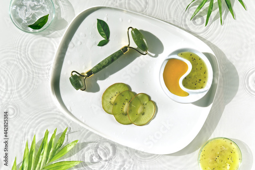 Home made cosmetic wth kiwi fruit. Self made skincare background. Jade stone face roller. In yang symbol plate filled with honey and homogenized kiwi. Anti aging, anti wrinkle. Sunlight, long shadows. photo