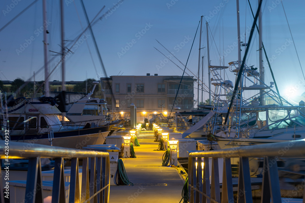 Marina dock and fishing boats with tall masts, on a foggy, hazy night. A ramp with railing to dock is in foreground and buildings in the distance.