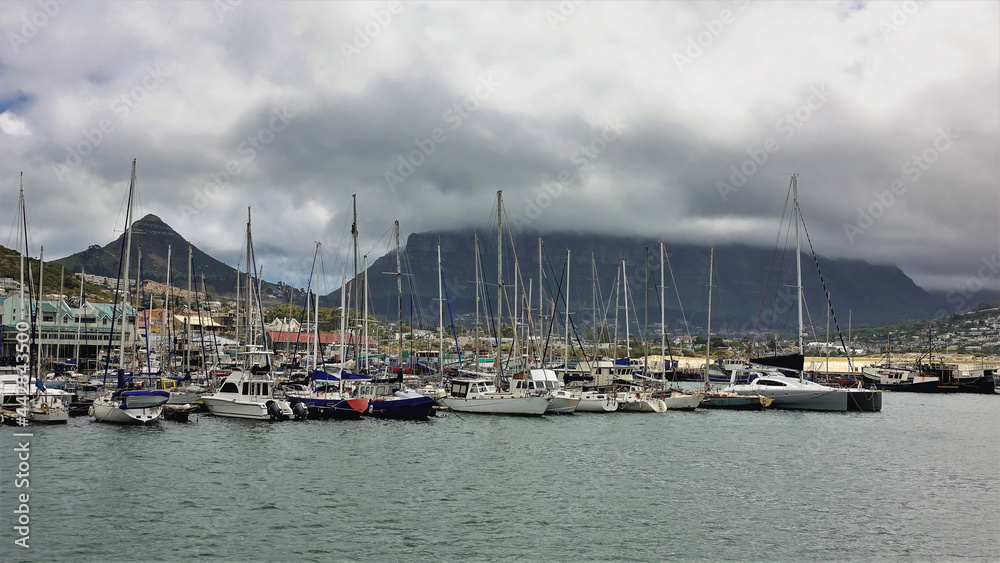 There are many sailing boats in Cape Town Bay. High masts against the backdrop of Table Mountain and Lion's Head Hill. Low dark clouds in the sky. South Africa