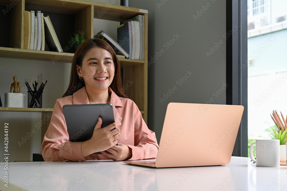 Fototapeta premium Smiling young woman holding digital tablet and sitting at office desk.