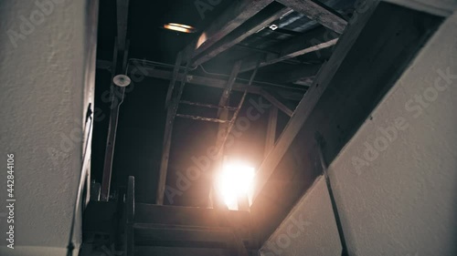 bottom view of an open roof hatch with old and worn staircase and bright orange lens flare shining through the skylight photo