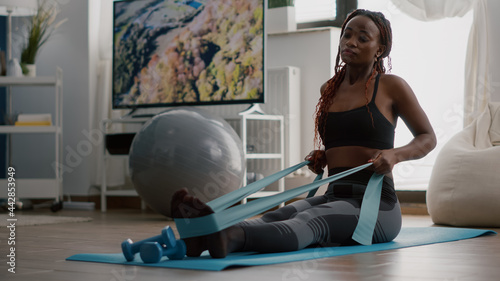 Athlete with black skin in sportswear exercising body muscle using fitness elastic enjoying healthy lifestyle sitting on yoga map in living room. Fit woman working at wellness warming before workout photo