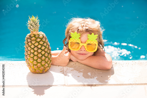 Little boy playing in outdoor swimming pool in blue water on summer vacation on tropical beach. Child learning to swim in pool of luxury resort. Summer pineapple fruit.