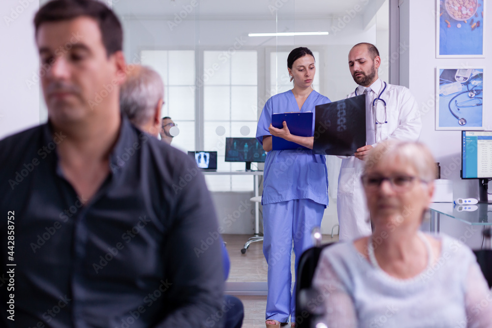 Obraz premium Specialist doctor holding patient x-ray explaining disease diagnosis to nurse while standing in hospital waiting area. Disabled senior woman in wheelchair waiting for medical examination