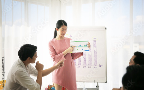 Smiling millennial female coach or presenter talk interact with multiracial colleagues at meeting, happy young woman tutor make flip chart whiteboard presentation at office briefing