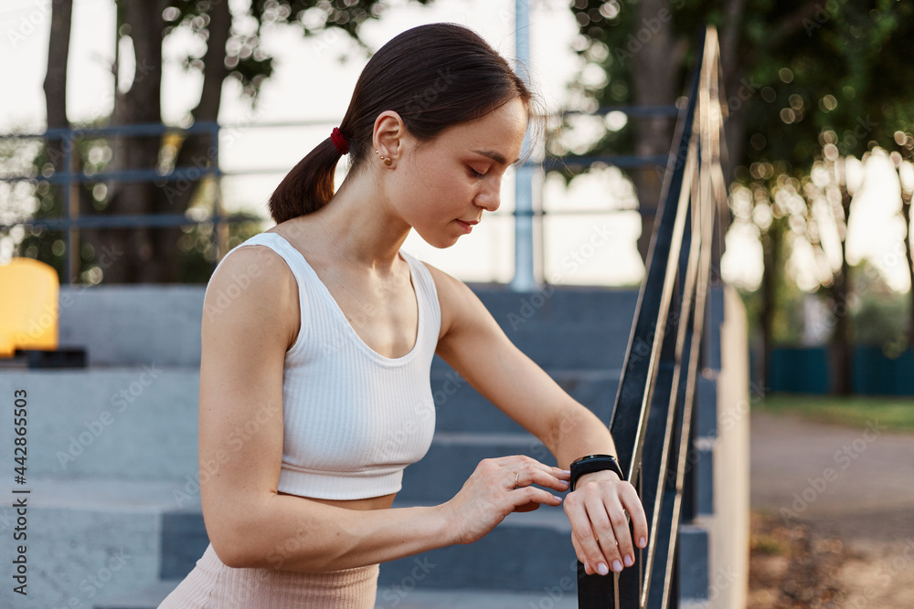 Healthy lifestyle and sport in summer. Side view of dark haired woman happy with result of jogging and looks at fitness tracker with concentrated look.