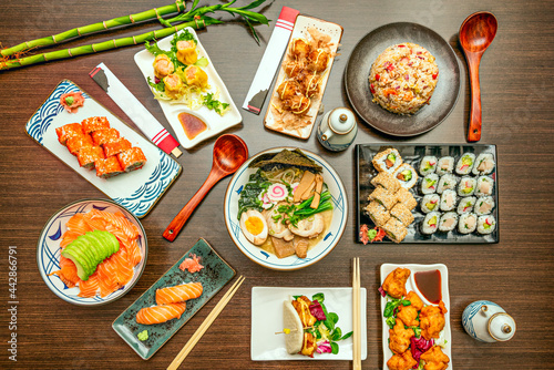 Top view image of Asian dishes featuring salmon sashimi with avocado, fish and vegetable ramen, assorted trays of uramaki and maki, California roll, soy sauce, bao bread