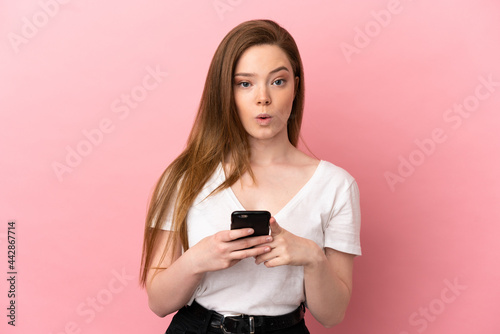 Teenager girl over isolated pink background looking at the camera while using the mobile with surprised expression
