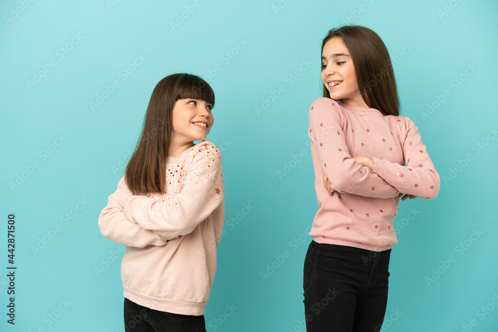 Little sisters girls isolated on blue background looking over the shoulder with a smile Little sisters girls isolated on blue background looking over the shoulder with a smile