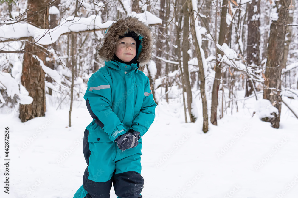 A boy in a turquoise overalls with a hood walks in a snowy forest and smiles. People, lifestyle concept