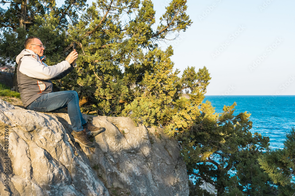 A Caucasian pensioner, age 60, photographs the sea on a smartphone while sitting on a mountain among mountain trees during a tourist trip at sunset