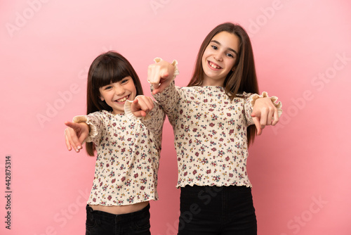 Little sisters girls isolated on pink background points finger at you while smiling