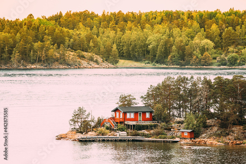 Sweden. Many Beautiful Red Swedish Wooden Log Cabin House On Rocky Island Coast In Summer. Lake Or River Landscape