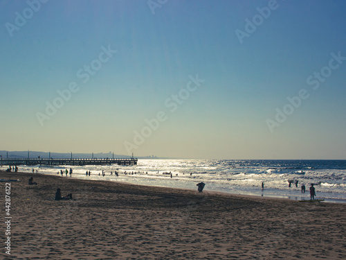 A beach filled with beach goers  playing on the sand and in the water  with a pier in the background and the sun reflecting off the water