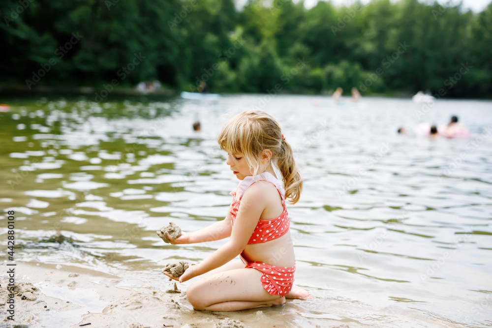 Little blond preschool girl having fun with playing with sand on lake on summer day, outdoors. Happy child learning swimming. Active leisure with kids on vacations. Danger on domestic lakes Little blond preschool girl having fun with playing with sand on lake on summer day, outdoors. Happy child learning swimming. Active leisure with kids on vacations. Danger on domestic lakes