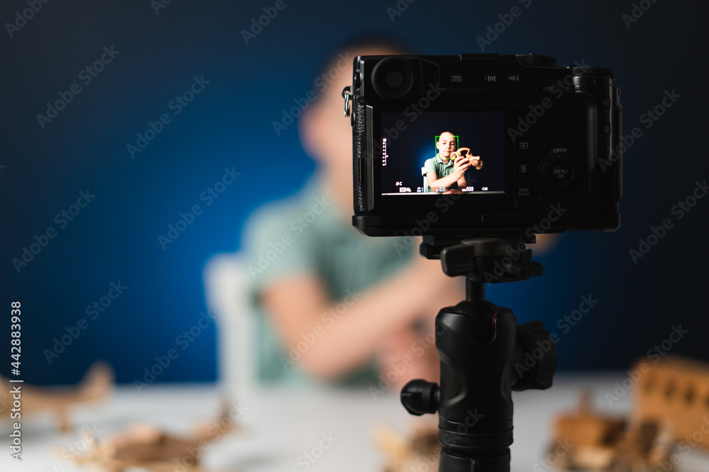 Kid blogger filming video on camera indoors with blue light in background. Online reviewing. Promoting eco wooden toys. Kid recording his gameplay for vlog.
