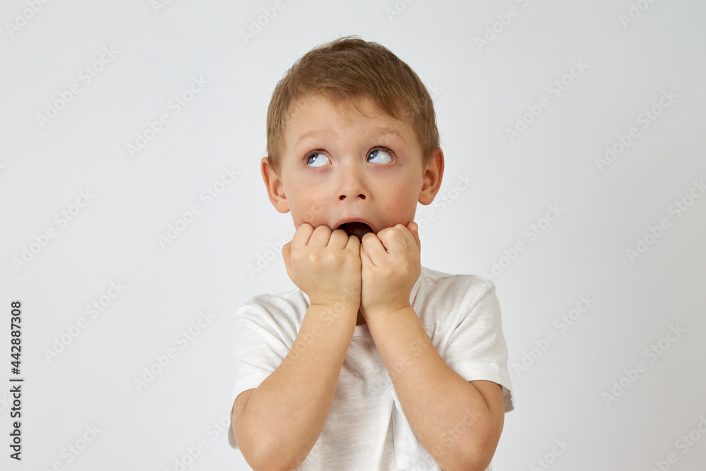 Child with horror looks up biting his hands in fear on a white background. Emotion of fear on the face of the boy with blue eyes Child with horror looks up biting his hands in fear on a white background. Emotion of fear on the face of the boy with blue eyes