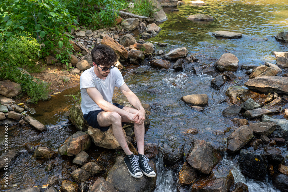 Sad young man sits on stepping stones in flowing stream looking down Sad young man sits on stepping stones in flowing stream looking down
