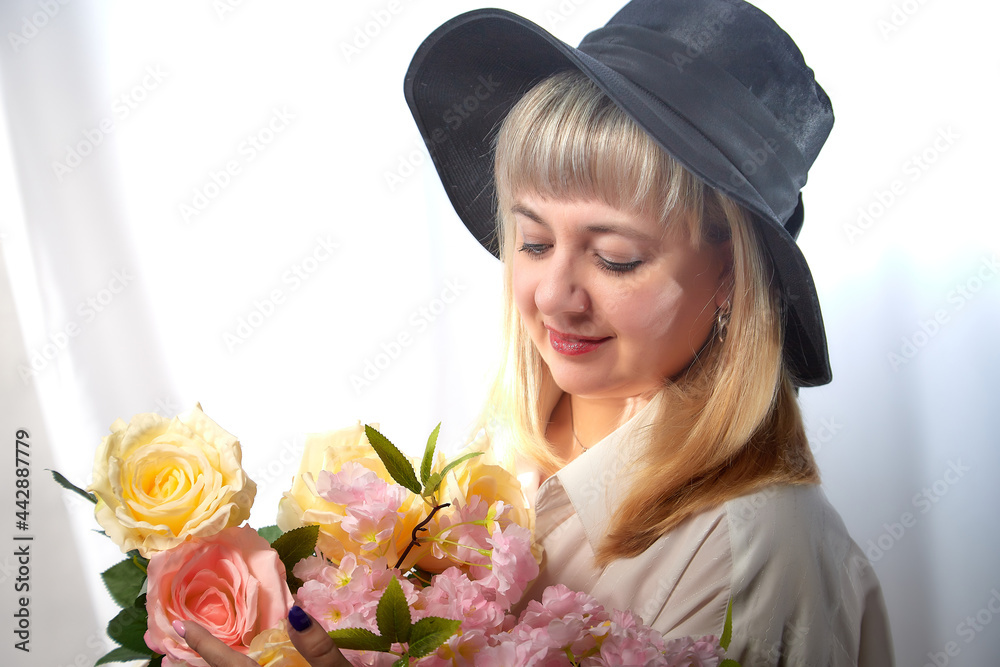 Happy beautiful woman with long blond hair and in black hat posing with flowers in studio on white background