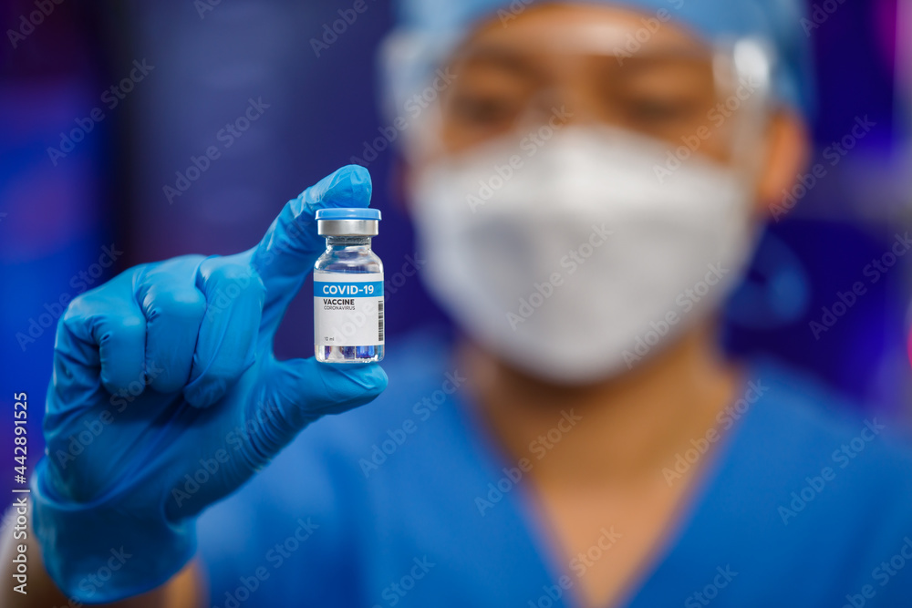 Female medical people wearing a hygiene face mask and protective glass staying in laboratory room, holding Covid-19 vaccine bottle with lab equipment in blur background Female medical people wearing a hygiene face mask and protective glass staying in laboratory room, holding Covid-19 vaccine bottle with lab equipment in blur background