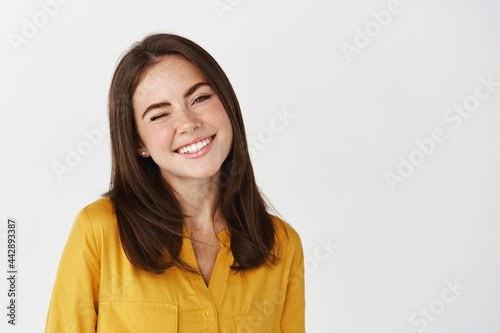 Close-up of happy young woman winking at camera, smiling joyful, standing over white background