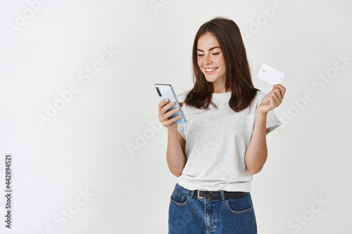 Online shopping. Young happy woman paying with smartphone and plastic credit card, standing over white background