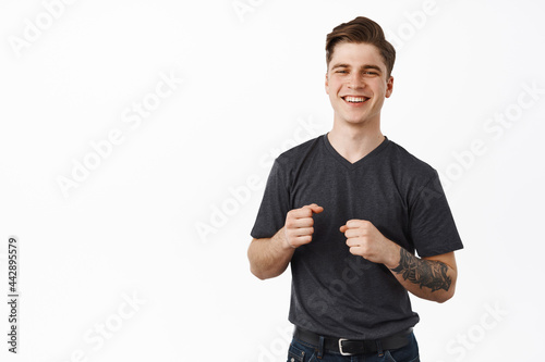 Image of handsome smiling guy awkward dancing. Young man laughing and holding fists clenched for good luck, standing against white background, holding in hands