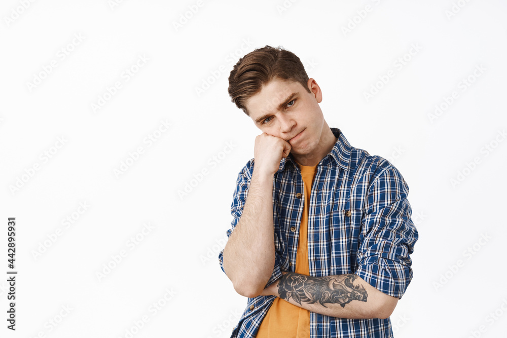 Teenage boy sulking, looks angry or annoyed, lean on fist and pouting upset, stands moody and sad against white background Teenage boy sulking, looks angry or annoyed, lean on fist and pouting upset, stands moody and sad against white background