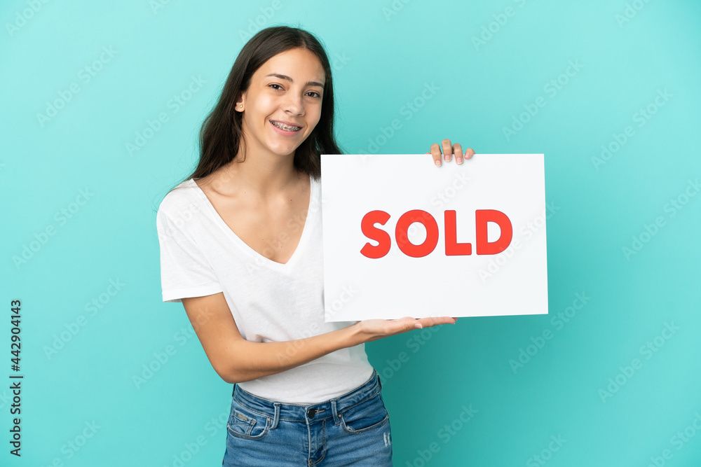 Young French woman isolated on blue background holding a placard with text SOLD with happy expression Young French woman isolated on blue background holding a placard with text SOLD with happy expression