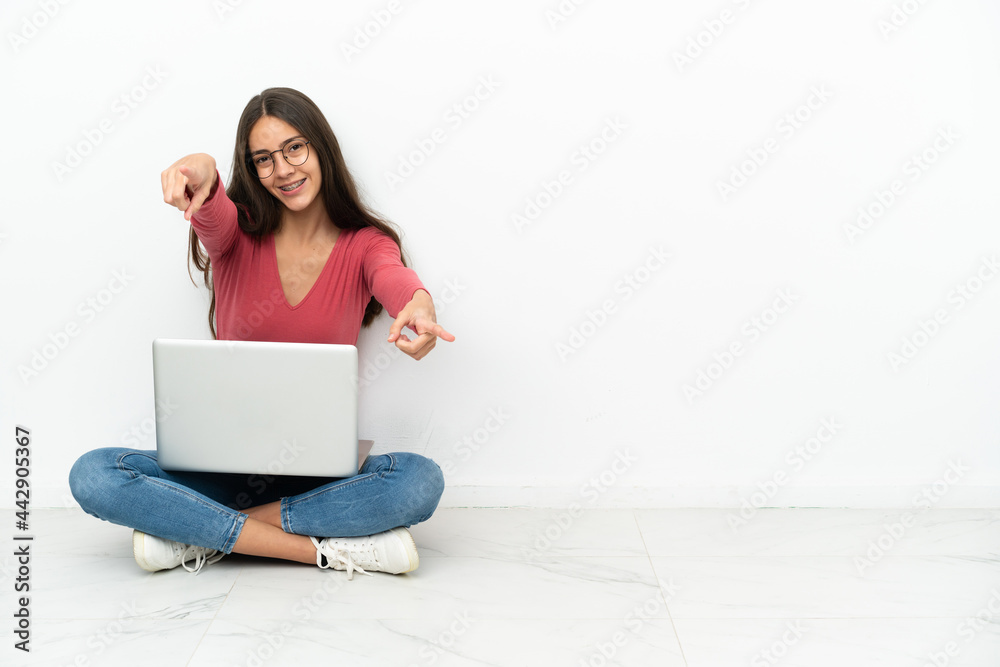 Young French girl sitting on the floor with her laptop points finger at you while smiling Young French girl sitting on the floor with her laptop points finger at you while smiling