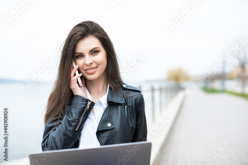 A young pretty girl, dressed in a leather jacket and jeans, spends time alone on the river bank and works with a laptop in hand. The girl looks at the laptop screen and talks on the phone