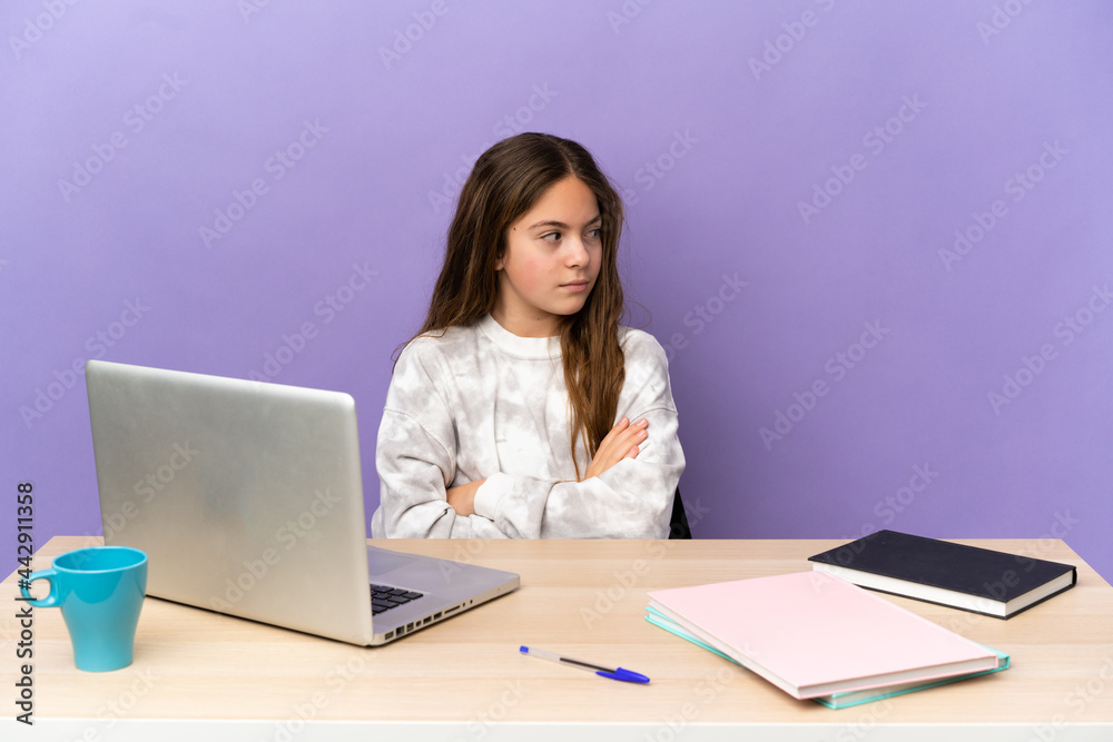 Little student girl in a workplace with a laptop isolated on purple background keeping the arms crossed Little student girl in a workplace with a laptop isolated on purple background keeping the arms crossed