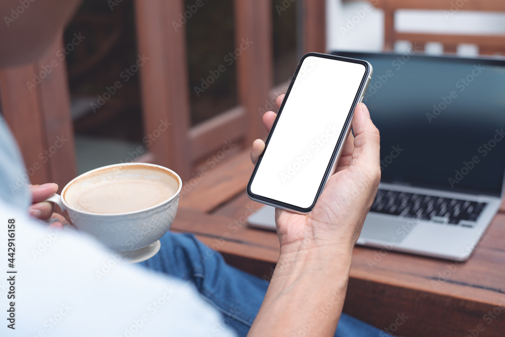 Mockup image blank white screen cell phone. Man hand holding, using mobile and drinking coffee during working on laptop in coffee shop