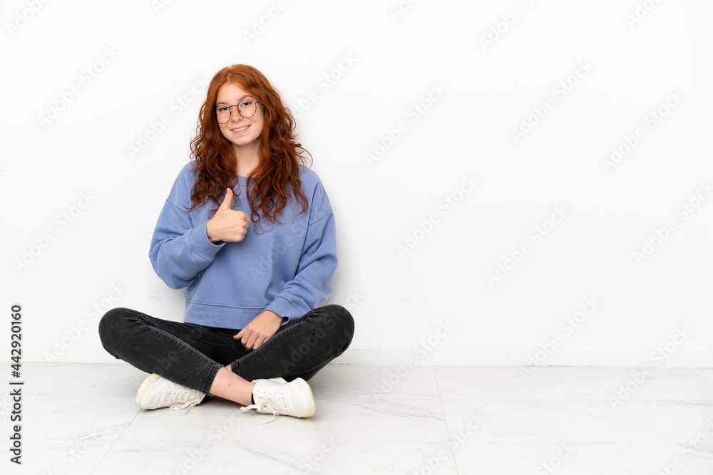 Teenager redhead girl sitting on the floor isolated on white background giving a thumbs up gesture Teenager redhead girl sitting on the floor isolated on white background giving a thumbs up gesture