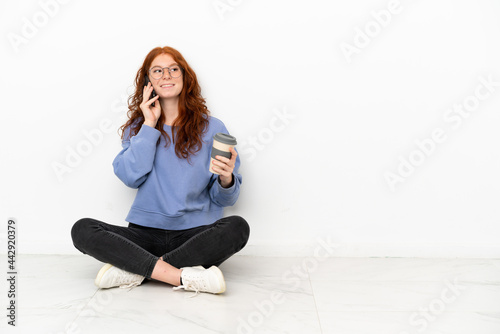 Teenager redhead girl sitting on the floor isolated on white background holding coffee to take away and a mobile